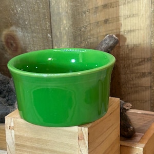 May include: A bright green ceramic bowl with a rounded shape. The bowl has a glossy finish and sits on a wooden block. The background features wooden planks and a small wooden box.
