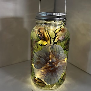 May include: A decorative glass jar lantern with a floral design. The jar features a painted flower and leaf pattern in shades of purple, green, and gold. The jar is lit with small fairy lights and has a metal handle.