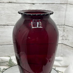 May include: A tall, ruby-red glass vase with a flared, scalloped rim. The vase has a smooth, glossy finish and a classic, elongated shape. The vase is likely used for decorative purposes.