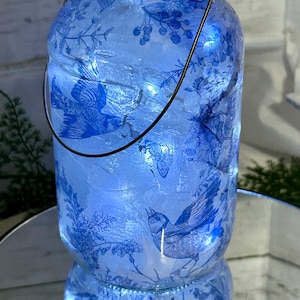 May include: A decorative glass jar with a blue and white bird and floral design. The jar has a metal handle and lid, and is illuminated with small white lights. The jar is sitting on a reflective surface.