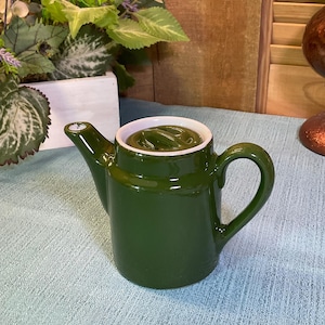 May include: A small, glossy, dark olive green ceramic teapot with a matching lid, a curved handle, and a short, angled spout. The interior rim of the teapot is white, and the exterior surface features a smooth, reflective finish throughout the body.