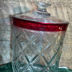 May include: A clear glass jar with a diamond pattern and a red rim. The jar has a clear glass lid with a knob. The jar is sitting on a green surface.
