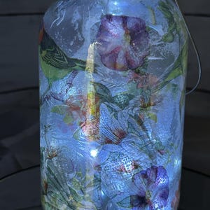 May include: A glass jar with a wire handle, decorated with a floral pattern and lit from within by white fairy lights. The jar is filled with a blue liquid.