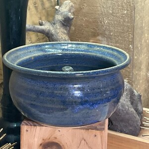 May include: A round, deep ceramic bowl with a glossy, mottled blue glaze. The bowl features a wide, slightly flared rim and a small, centered knob at the bottom of the interior. The exterior surface shows subtle horizontal ridges along the curved body.