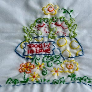May include: Embroidered white fabric with two green frogs sitting on a yellow mushroom with the words "badly in love" on a sign. The mushroom is surrounded by green leaves and yellow flowers.