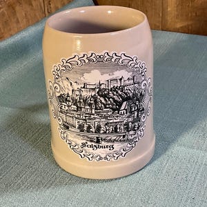 May include: A beige ceramic beer mug featuring a black and white illustration of the city of Salzburg, Austria. The detailed graphic shows buildings, a fortress on a hill, and a bridge, all enclosed within an ornate, decorative circular border.