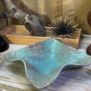 May include: A square ceramic bowl with a wavy, undulating rim. The bowl features a textured, speckled glaze in shades of turquoise and teal, with patches of reddish-brown oxidation visible along the edges and across the interior surface.