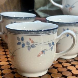 May include: Set of four cream-colored ceramic mugs. Each mug features a blue rim and a delicate floral pattern with blue leaves and pink flowers. The mugs have a curved handle and are suitable for hot beverages.