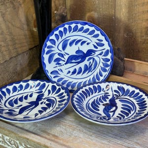 May include: Three round ceramic plates featuring a hand-painted cobalt blue bird motif surrounded by a border of blue leaf-like brushstrokes on a white background. The plates are arranged with one upright and two positioned in the foreground.