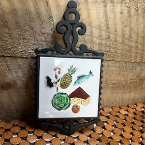 May include: Black cast iron trivet with a decorative scrolled handle and frame. The white tile insert features colorful illustrations of a rooster, pineapple, fish, cheese, cabbage, and tomato. A kitchen decor item.