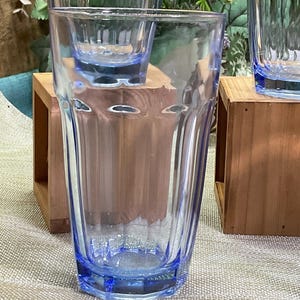May include: A clear light blue glass tumbler featuring a faceted, vertical ribbed design. The drinking glass has a wide rim that tapers down to a narrower, hexagonal base, showcasing a transparent construction with a distinct blue tint throughout.