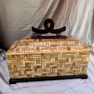 May include: A rectangular storage box featuring a light tan and brown woven-look pattern on its exterior. The box has a dark brown base and a matching dark brown lid topped with a decorative, curved metal handle featuring two small spheres.