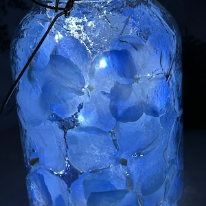 May include: A glass jar with a textured surface filled with blue flower petals and white lights. The jar has a metal lid.