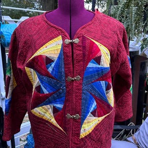 May include: A quilted jacket in deep red with a starburst design in shades of blue, yellow, and red. The jacket has a V-neck and is fastened with three ornate clasps. The sleeves are three-quarter length.