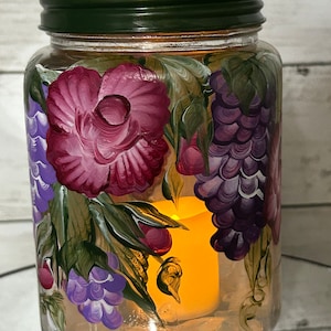 May include: A glass jar with a green lid, hand-painted with purple grapes, pink flowers, and green leaves. A candle is inside the jar, casting a warm glow.