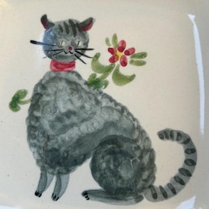 May include: A gray cat with a red collar and a green flower behind it is painted on a white ceramic square dish.