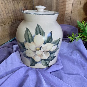 May include: A cream-colored ceramic canister with a lid, featuring a hand-painted illustration of a large white magnolia flower with dark green leaves on the front. The rim of the canister and the lid have a speckled, dark gray textured edge.