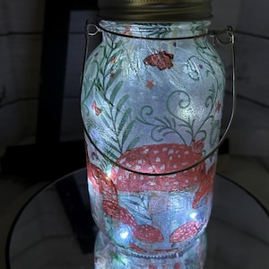 May include: A glass jar with a metal handle and a white lid. The jar is decorated with a mushroom and butterfly design. The jar is lit from within with white fairy lights.