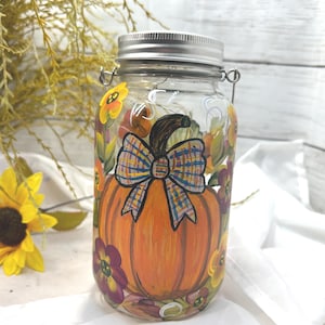 May include: A clear glass jar with a silver lid, painted with an orange pumpkin and a plaid bow. The jar is decorated with yellow and purple flowers, perfect for fall decor.