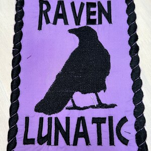 May include: Purple fabric patch with black embroidered text and a black silhouette of a raven. The text reads "RAVEN LUNATIC".