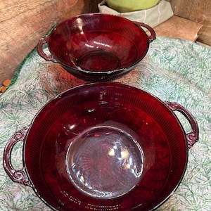 May include: Two ruby red glass bowls with handles. The bowls have a ribbed design on the sides and a smooth, circular base. The handles have a decorative, scrolled design. The bowls are likely vintage or antique.