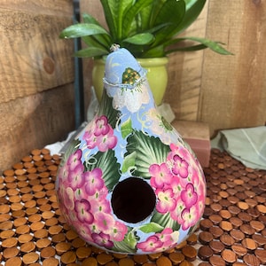 May include: A hand-painted gourd birdhouse with a light blue background and pink hydrangea flowers. The birdhouse has a round entry hole and a wire loop for hanging. The gourd is sitting on a wooden surface.