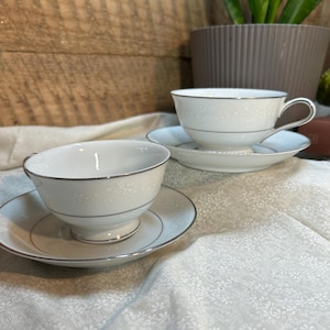 May include: Two white teacups and saucers with a delicate floral pattern and silver trim. The teacups are on a white patterned tablecloth. A plant in a brown pot is in the background.