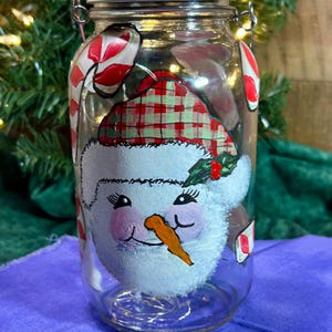 May include: A clear glass jar with a silver lid, decorated with a hand-painted snowman face and candy cane accents. The snowman has a plaid hat, rosy cheeks, and an orange carrot nose. The jar is lit with fairy lights.