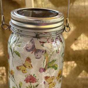 May include: A glass jar with a metal lid and a wire handle. The jar is decorated with a floral pattern and butterflies. The lid has a solar panel on top.