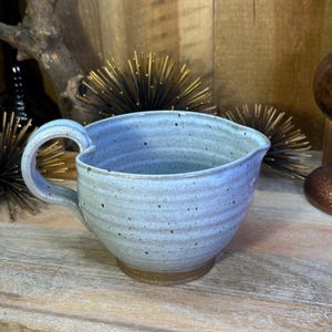 May include: A light blue ceramic pitcher with a speckled finish and horizontal ridges along the body. The pitcher features a single curved handle on the left side and a small pouring spout on the right side, with a dark brown unglazed base.