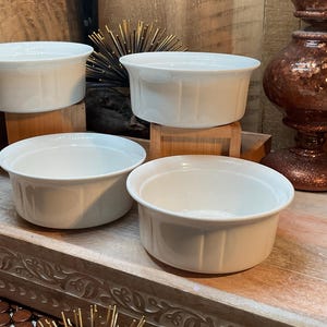 May include: Four white, round, ceramic ramekins with a slightly flared rim. The ramekins are arranged on a wooden surface. The background includes decorative items and a wooden wall. These ramekins are suitable for baking and serving individual portions.