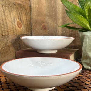 May include: Two white ceramic bowls with a tan rim and base. The bowls are set on a wooden surface, with a wooden background and a potted plant visible. The bowls have a simple, elegant design.