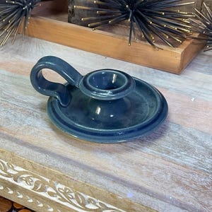May include: A dark blue ceramic candle holder with a glossy finish, featuring a circular base, a central raised socket for a taper candle, and a curved handle attached to the side for easy carrying, showcasing a smooth, uniform glazed surface.