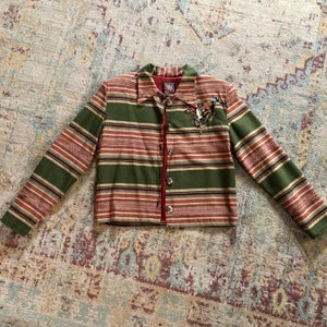 May include: A green, red, and brown striped jacket with a decorative pin on the front. The jacket has a collared neckline, long sleeves, and silver buttons. The interior lining is red.