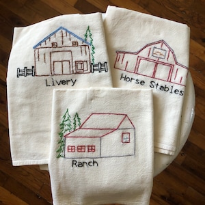 May include: Four off-white embroidered kitchen towels. Each towel features a different barn design with the words "Livery", "Horse Stables", and "Ranch" embroidered below the barn illustrations. The barns are in shades of brown, red, and blue.