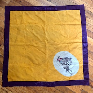May include: A square, yellow and purple bandana with text that reads "The Round-up", "Oregon", "Pendleton", and "Let Her Buck". The bandana has a purple border and a white oval with a horse and rider illustration.