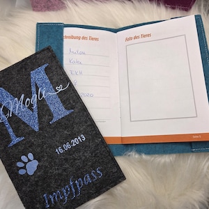 May include: A gray felt pet passport cover with a blue glitter "M" and the name "Mogli". The cover includes a paw print and the date "16.06.2013". An open teal and white booklet is visible, with the text "Foto des Tieres".