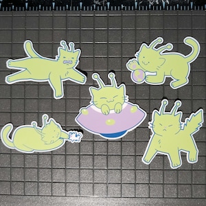 May include: A collection of five stickers featuring cartoon alien cats in various poses. The cats are light green with white outlines and antennae. One cat is in a purple and pink UFO. The stickers are on a black grid.