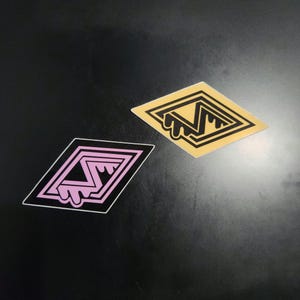 May include: Two diamond-shaped stickers. One is gold with a black design and dripping detail. The other is pink with a black border and a black design. Both stickers have a unique, stylized design.