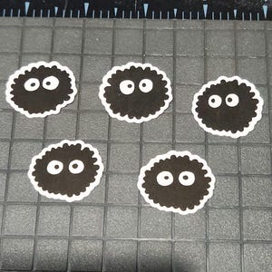 May include: Five black, round stickers with white, oval eyes and a white border. The stickers have a fuzzy, textured appearance and are arranged on a grey, grid-patterned surface.