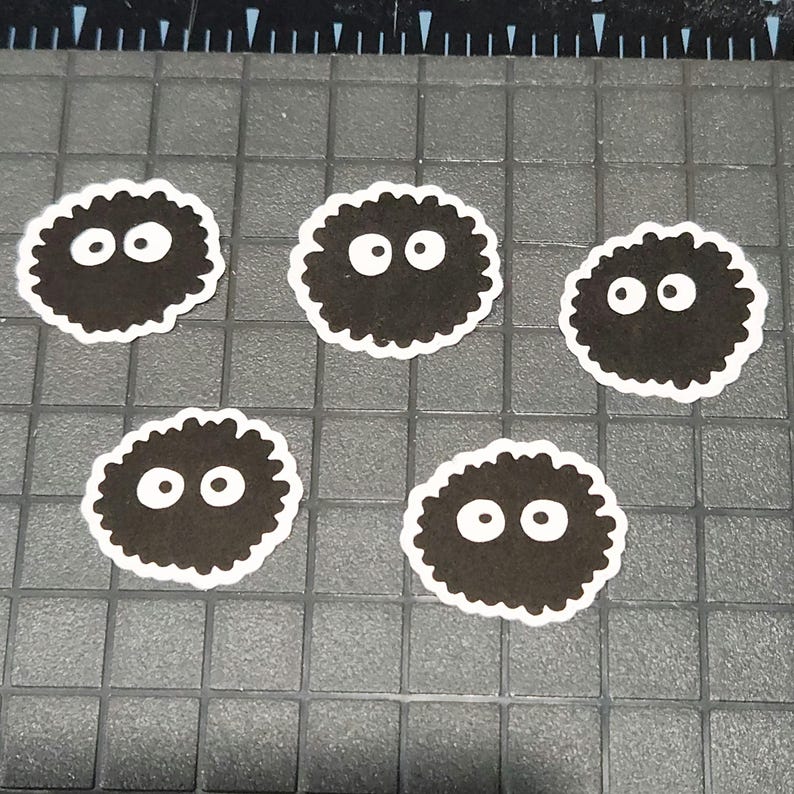 May include: Five black, round stickers with white, oval eyes and a white border. The stickers have a fuzzy, textured appearance and are arranged on a grey, grid-patterned surface.
