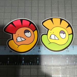 May include: Two cartoon stickers with a circular design. One is orange and red, the other is yellow and green. Both have a face with a white eye, a frown, and a black outline.