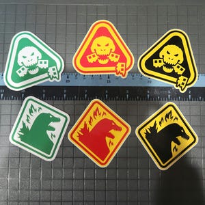 May include: A collection of six stickers featuring monster-themed designs. The stickers include triangular and square shapes with images of a skull and a monster, in green, red, yellow, and black colors. The stickers have white borders.