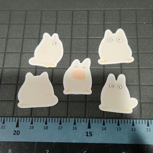 May include: Five white die-cut stickers depicting a cartoon character with rounded ears and a small tail. Some stickers have eyes, and one has a pink nose. The stickers have a yellow border and are arranged on a black grid.