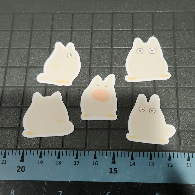 May include: Five white die-cut stickers depicting a cartoon character with rounded ears and a small tail. Some stickers have eyes, and one has a pink nose. The stickers have a yellow border and are arranged on a black grid.
