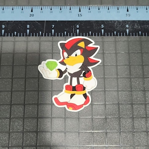 May include: A die-cut sticker featuring the character Shadow the Hedgehog from the Sonic the Hedgehog franchise. Shadow is depicted in his signature black, red, and white color scheme, holding a green Chaos Emerald. The sticker is on a gray surface with measurement markings.