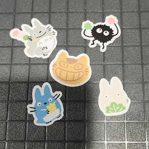 May include: Five cartoon stickers featuring characters from the animated film "My Neighbour Totoro." The stickers include a grey Totoro, a black soot sprite, a smiling orange cat, a blue Totoro, and a white bunny-like creature.