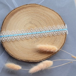 Puede incluir: Borde de encaje blanco con una cinta azul claro sobre una rodaja redonda de madera. Dos tallos de hierba seca beige est&aacute;n sobre la rodaja de madera.
