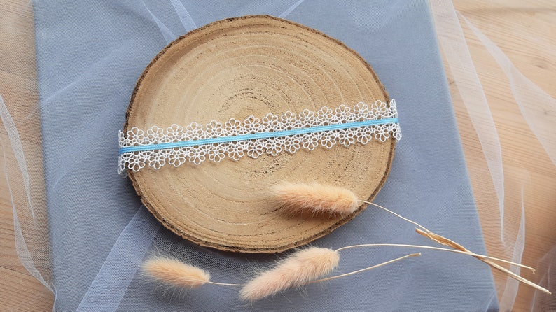 Puede incluir: Borde de encaje blanco con una cinta azul claro sobre una rodaja redonda de madera. Dos tallos de hierba seca beige est&aacute;n sobre la rodaja de madera.