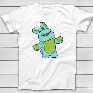 Puede incluir: Camiseta blanca con una ilustraci&oacute;n de dibujos animados de un personaje de conejo azul y verde con ojos rosados. El conejo tiene los brazos extendidos. El dise&ntilde;o est&aacute; centrado en la parte delantera de la camiseta.